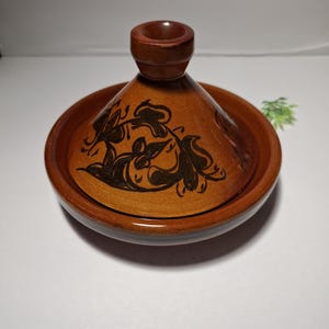 May include: A brown ceramic tagine pot with a conical lid and a round base. The lid features a dark floral design. The pot is designed for cooking and serving food. The tagine is a traditional North African cooking vessel.