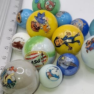 May include: A collection of colorful marbles with various cartoon character designs. The marbles are in shades of blue, yellow, green, and white. Some marbles feature images of cartoon characters, while others have logos. A ruler is in the background.