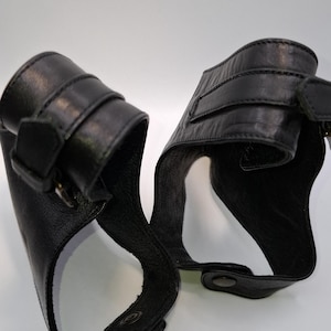 Puede incluir: Dos muñequeras de cuero negro con cierres de hebilla y correas ajustables. Las muñequeras tienen un diseño curvo y están sobre un fondo blanco. El cuero parece texturizado.