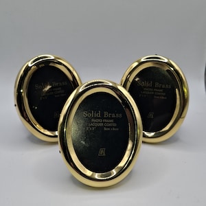 Könnte beinhalten: Drei goldfarbene, ovale Fotorahmen mit schwarzem Innenraum. Die Rahmen sind mit "Solid Brass" und "Photo Frame Lacquer Coated 2" x 3" 5cm x 8cm" beschriftet. Sie liegen auf einer weißen Oberfläche.