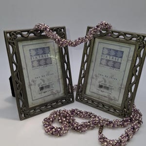 May include: Two decorative metal picture frames with ornate edges, connected by a beaded strand. The frames have a distressed, antique finish and hold printed inserts. The text "SIXTREES" and "two by three" are visible on the inserts. The beaded strand is a mix of purple and silver beads.