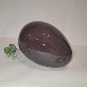 以下が含まれることがあります： 大きく光沢のある卵型の装飾品。表面は濃い紫の色合いで、茶色とグレーの微妙なアクセントがあります。小さな緑色の植物がオブジェクトの左側に配置され、白い背景に配置されています。