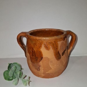 May include: A small, rustic, brown ceramic mug with two handles. The mug has a rounded shape and a textured surface with darker brown drips. A sprig of green foliage sits to the left of the mug.