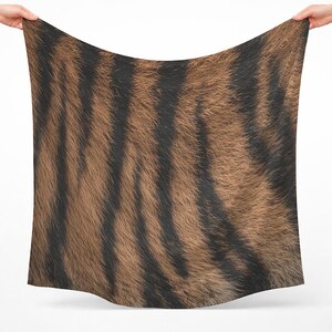 Puede incluir: Un pañuelo cuadrado con un estampado realista de piel de tigre que presenta un patrón de rayas verticales de color negro oscuro sobre un fondo marrón dorado texturizado. La tela muestra una apariencia fina y vellosa en toda su superficie.