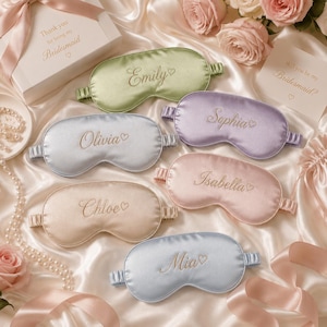 Op de afbeelding: Zes satijnen slaapmaskers in saliegroen, lavendel, lichtblauw, oudroze, champagne en maagdenpalm, elk geborduurd met een naam en een klein hartje. De namen zijn Emily, Sophia, Olivia, Isabella, Chloe en Mia, voorzien van elastische banden.