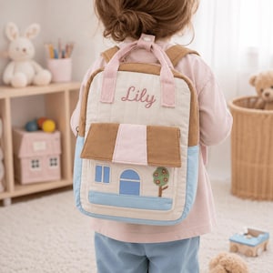 Puede incluir: Una mochila acolchada con diseño de casa, techo marrón, base azul claro y una aplicación de árbol. La bolsa tiene asas superiores rosas, correas ajustables y el nombre Lily bordado en letra cursiva rosa sobre la sección superior color crema.