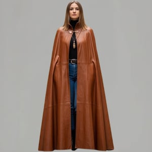 May include: A full-length, brown leather cape with a high collar and a decorative clasp. The cape is open in the front, revealing a black turtleneck, blue jeans, and a belt. The cape has a flowing, elegant design.