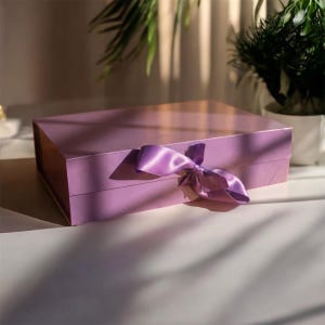 Può includere: Una confezione regalo lilla con un nastro di raso abbinato legato a fiocco. La scatola rettangolare è su una superficie bianca, con ombre di piante che aggiungono profondità. La scatola è chiusa e pronta per essere regalata.