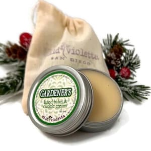 May include: A small tin of hand balm and cuticle cream with a green label that reads "GARDENER'S hand balm & cuticle cream" and "wild violetta". The tin is sitting on a white surface next to a small white drawstring bag.