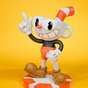 May include: A cartoon figurine featuring a white cup-shaped head, black arms, and orange shorts. The character is giving a peace sign and stands on a red and white striped platform. The background is a solid yellow.