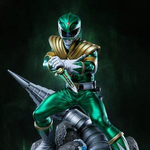 May include: A detailed statue of the Green Ranger from Power Rangers. The figure is in a dynamic pose, clad in a green and gold suit with a helmet. The statue is mounted on a rocky base with water effects and a large mechanical element.