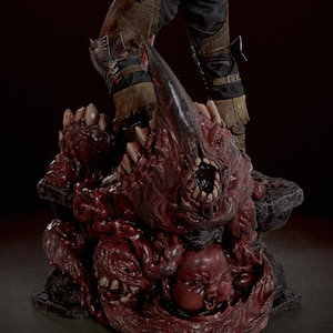 May include: A detailed collectible figurine base featuring a cluster of grotesque, fleshy, dark red organic masses with sharp teeth and a small, distorted face. The base supports the lower legs of a figure wearing brown leather and black trousers.