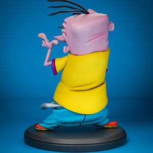 May include: A figurine of a cartoon character, viewed from the back, with a pink head, black hair, and a yellow shirt. The character is wearing blue pants and red shoes. The figurine is on a black circular base against a blue background.
