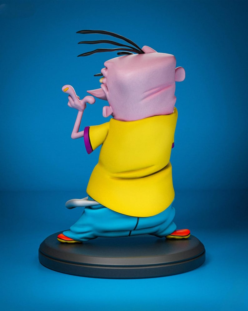 May include: A figurine of a cartoon character, viewed from the back, with a pink head, black hair, and a yellow shirt. The character is wearing blue pants and red shoes. The figurine is on a black circular base against a blue background.