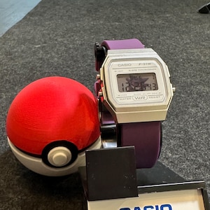 Puede incluir: Un reloj digital Casio F-91W plateado con una correa morada se muestra junto a una Pokeball roja y blanca. La esfera del reloj muestra la hora. Una caja de exhibición Casio está en primer plano.