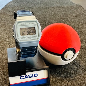 Puede incluir: Un reloj digital plateado Casio F-91W con una pantalla negra que muestra la hora, junto a una Pokeball roja, blanca y negra. El reloj está sobre un soporte negro con el logotipo de Casio en blanco y azul.