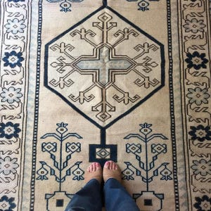 May include: A rectangular area rug featuring a cream-colored background with a large central dark blue geometric diamond motif containing a cross shape. The rug includes symmetrical floral patterns in shades of blue and dark blue along the borders.