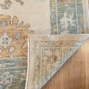 May include: Close-up of a beige and blue patterned rug with a folded corner. The rug features a repeating floral design in shades of beige, blue, and gold. The rug's texture is visible, and the corner reveals the rug's backing.