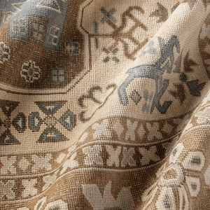 May include: A woven textile rug featuring a complex pattern of geometric shapes and stylised floral motifs. The colour palette consists of soft beige, warm brown, and muted blue tones, with a textured surface that shows folds and depth.