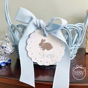 May include: A light blue wicker Easter basket with a matching ribbon and a decorative tag. The tag features a brown embroidered bunny and the name "Shep". The basket is on a wooden surface.