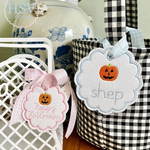 Może przedstawiać: Dwie wykończone w ząbki materiałowe zawieszki z wyhaftowanymi pomarańczowymi dyniami. Jedna zawieszka zawiera napis "Happy Halloween" w kolorze różowym i jest przewiązana różową wstążką w kratkę. Druga zawiera imię "shep" w kolorze niebieskim i jest przymocowana jasnoniebieską wstążką do czarno-białego koszyka z materiału w kratkę.