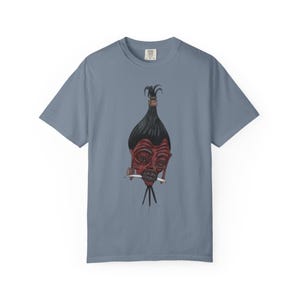 May include: A blue-grey t-shirt featuring a graphic of a red face with black hair and a knife in its mouth. The artwork has a unique, artistic style. The shirt is made of a soft material.