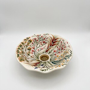 May include: Cream-colored ceramic sink with a floral design. The sink features a scalloped edge and a central drain with a gold-colored cover. The floral pattern includes red, pink, and green botanical elements.