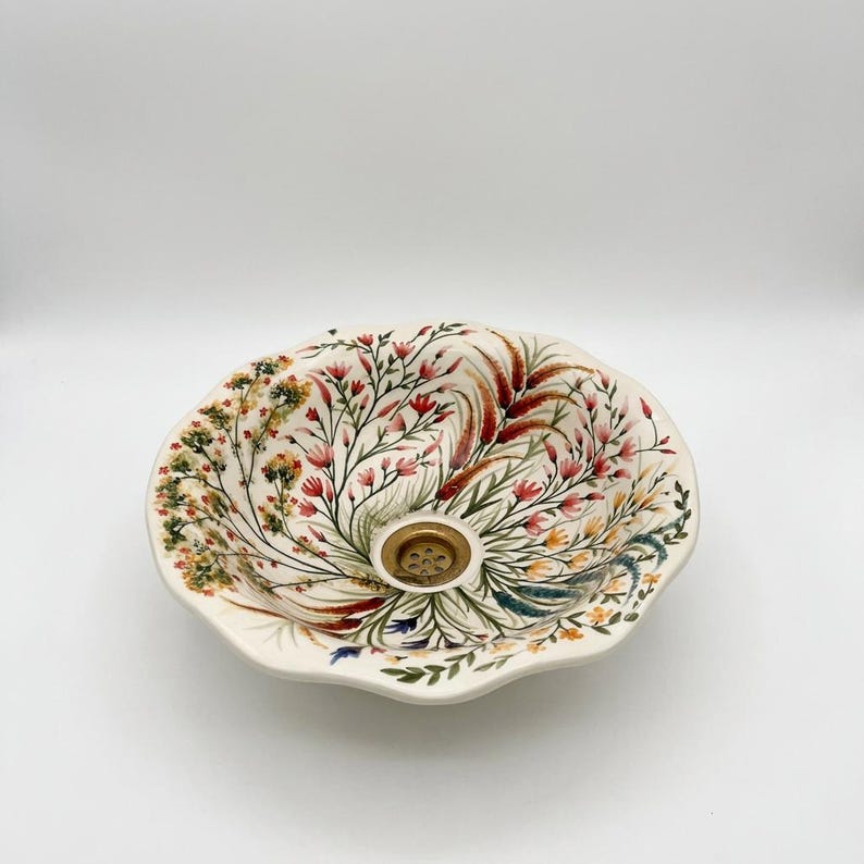 May include: Cream-colored ceramic sink with a floral design. The sink features a scalloped edge and a central drain with a gold-colored cover. The floral pattern includes red, pink, and green botanical elements.
