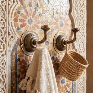 Pode incluir: Dois ganchos de parede de latão antigo com um design circular decorativo são montados em uma parede de azulejos de mosaico. Uma toalha bege com borlas pende de um gancho e uma pequena cesta tecida do outro.
