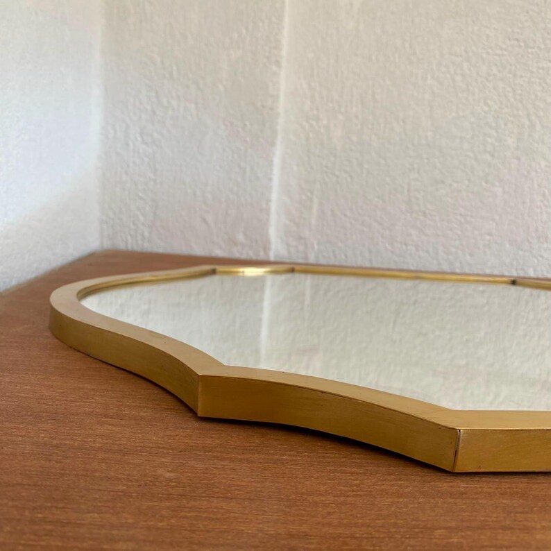 May include: A wall mirror featuring a reflective glass surface encased in a smooth, metallic gold frame with a distinct scalloped or quatrefoil-inspired shape. The frame has a flat, wide edge and a clean, polished finish that catches the light.