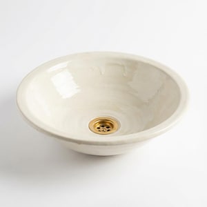 Puede incluir: Lavabo de cerámica blanco roto con forma redonda. El lavabo tiene un acabado brillante y un desagüe de latón. El diseño es sencillo y moderno, adecuado para un baño.