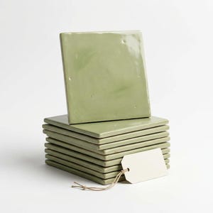 May include: A stack of eight square, sage green ceramic tiles with a glossy finish. The tiles have slightly uneven, organic edges and a smooth surface. A blank, rectangular white paper tag is attached to the stack with a thin brown string.