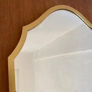May include: A wall mirror featuring a smooth, gold-toned metal frame with a distinct arched top and curved sides. The reflective glass surface shows a clear view of a white interior wall, while the frame edges are finished in a metallic gold.