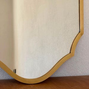 May include: A wall mirror featuring a reflective glass surface and a smooth, metallic gold-toned frame. The frame has a distinct curved, scalloped edge design that follows the contour of the mirror, creating a clean and defined border.