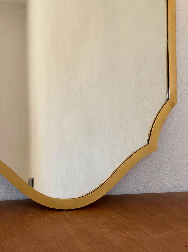 May include: A wall mirror featuring a reflective glass surface and a smooth, metallic gold-toned frame. The frame has a distinct curved, scalloped edge design that follows the contour of the mirror, creating a clean and defined border.