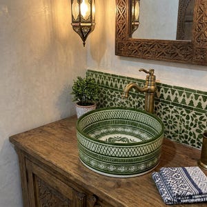 Puede incluir: Un lavabo de cerámica redondo con intrincados patrones geométricos verdes y blancos en el exterior e interior. El lavabo está colocado sobre un mueble de madera junto a un grifo de latón, con un salpicadero de azulejos verdes y una planta pequeña.