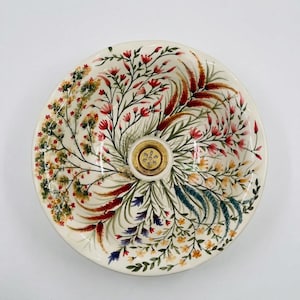 May include: A round, off-white ceramic decorative bowl with a floral design. The bowl features a central gold-colored fixture and is adorned with colorful hand-painted flowers and foliage in shades of red, green, yellow, and blue. The rim is slightly raised.
