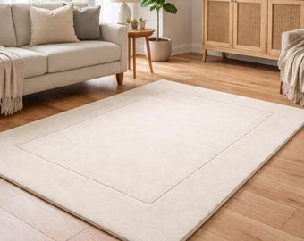 Peut inclure: Un tapis rectangulaire crème avec un bord subtil, posé sur un parquet. Le tapis est au centre d'un salon, avec un canapé, une table d'appoint et un meuble visibles en arrière-plan.