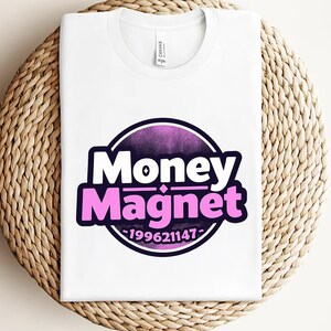 May include: White t-shirt with a circular graphic that reads "Money Magnet" in white and pink lettering, set against a purple gradient background. The number "199621147" is below the text. The shirt is folded on a woven, circular placemat.