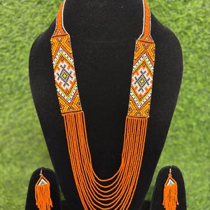 Puede incluir: Un collar de cuentas naranja y un conjunto de pendientes a juego exhibidos en un maniquí negro. El collar presenta un diseño geométrico en blanco, amarillo y negro. Los pendientes tienen una parte superior triangular y flecos de cuentas.