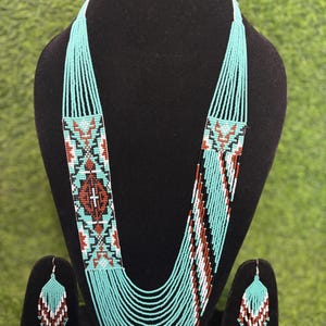 May include: A turquoise beaded necklace and matching earrings set. The necklace has a rectangular pendant with a geometric pattern in red, black, and white. The earrings have a similar design. The necklace and earrings are displayed on a black stand.