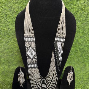 May include: A beaded necklace and matching earrings set. The necklace has a geometric design in black, white, and silver beads. The earrings have a fringe design. The jewellery is displayed on a black mannequin against a green background.