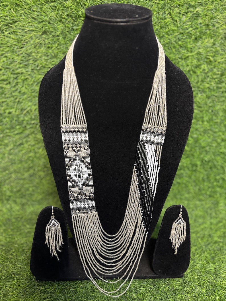 May include: A beaded necklace and matching earrings set. The necklace features a geometric design in black, white, and silver beads. The earrings have a fringe design. The jewelry is displayed on a black mannequin against a green background.