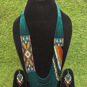 Puede incluir: Un collar de cuentas de múltiples hilos y un par de pendientes a juego. Las joyas presentan un patrón geométrico en cuentas de vidrio color verde azulado, naranja, blanco y negro. El collar tiene una sección tejida plana y ancha.