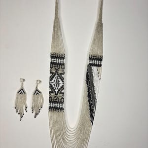 May include: A beaded necklace and matching earrings set. The necklace features a geometric pattern in black, white, and gray beads. The earrings have a fringe design with black and silver beads. The jewelry set is displayed on a white background.