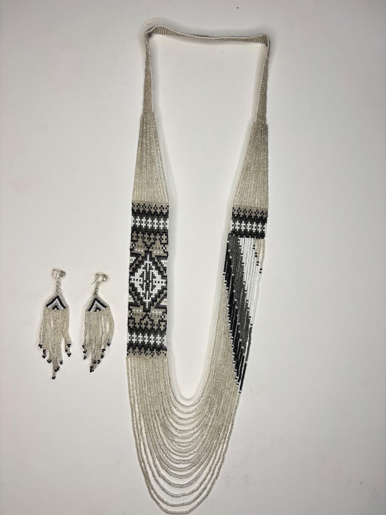 May include: A beaded necklace and matching earrings set. The necklace features a geometric pattern in black, white, and gray beads. The earrings have a fringe design with black and silver beads. The jewelry set is displayed on a white background.