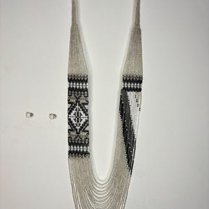 May include: A long, beaded necklace with a geometric design. The necklace features silver beads and sections of black, white, and gray beads. The design includes a diamond pattern and vertical stripes. The necklace hangs on a white background.