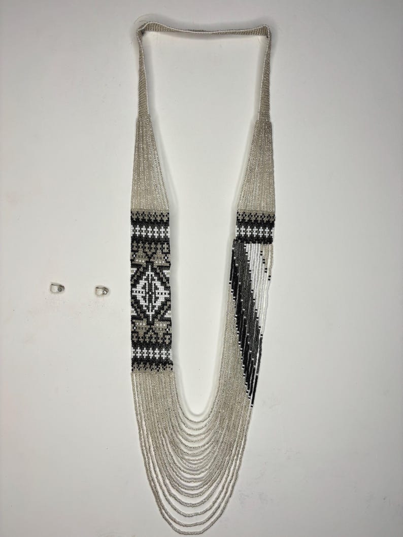 May include: A long, beaded necklace with a geometric design. The necklace features silver beads and sections of black, white, and gray beads. The design includes a diamond pattern and vertical stripes. The necklace hangs on a white background.