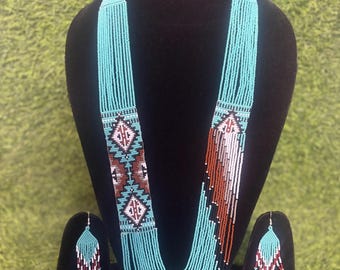 May include: A turquoise beaded necklace and matching earrings set. The necklace features a geometric pattern in brown, white, and black. The earrings have a similar design. The jewelry is displayed on a black mannequin against a green background.