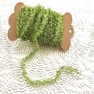 May include: A spool of lime green metallic tinsel trim wrapped around a brown cardboard bobbin. The trim has a fuzzy, reflective texture and a single strand extends from the spool, showcasing the vibrant green color and shimmering finish.